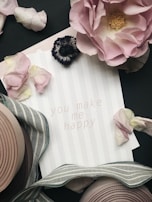 A greeting card with the message 'you make me happy' is surrounded by pink and white flower petals and a striped ribbon. A large pink rose and a dark flower are positioned at the top of the image, adding a romantic and elegant touch. The background features a subtle striped pattern.