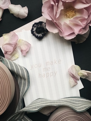 A greeting card with the message 'you make me happy' is surrounded by pink and white flower petals and a striped ribbon. A large pink rose and a dark flower are positioned at the top of the image, adding a romantic and elegant touch. The background features a subtle striped pattern.