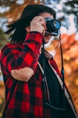 A person wearing a red and black checkered shirt is holding a camera up to their face, capturing a photo. The background features blurred autumn foliage with golden and orange leaves.