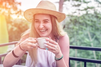 A woman with long blonde hair is laughing heartily while holding a cup of coffee. She is wearing a straw hat, a sleeveless white top, and several pieces of jewelry including rings, a bracelet, and a necklace. The background features lush green vegetation and a blurred railing, suggesting she is outdoors, possibly on a patio or balcony.