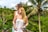A couple stands on a wooden platform surrounded by lush green tropical foliage, including tall palm trees. The woman, wearing a white dress, smiles while looking to the side, and the man, in a sleeveless shirt, stands behind her. The setting suggests a warm, exotic location.