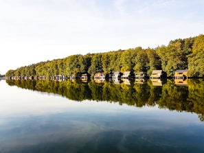 A serene lake with calm water reflecting a row of small wooden cabins and lush, green trees along the shoreline. The sky above is clear with a light cloud cover.