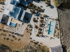 An aerial view of a modern property featuring a rectangular swimming pool surrounded by lounge chairs and a variety of vegetation. The building is white with a flat roof and multiple levels. The surrounding landscape is a mix of paved pathways, sandy areas, and scattered shrubs.