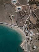 An aerial view of a coastal area with clear greenish-blue water, a sandy shore, and intersecting roads. The landscape includes fields with houses scattered around. There are patches of vegetation, possibly trees or shrubs, and cultivated land. The layout appears to be semi-rural with some residential buildings near the coastline with a few small piers extending into the water.