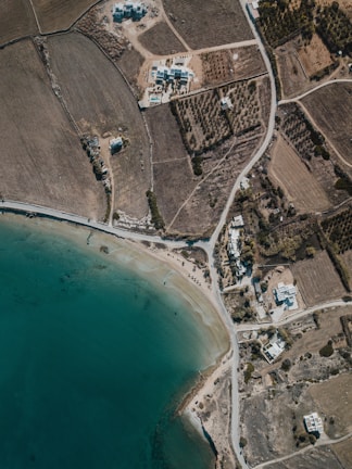An aerial view of a coastal area with clear greenish-blue water, a sandy shore, and intersecting roads. The landscape includes fields with houses scattered around. There are patches of vegetation, possibly trees or shrubs, and cultivated land. The layout appears to be semi-rural with some residential buildings near the coastline with a few small piers extending into the water.