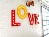 Large letters spelling out the word 'LOVE' are mounted on a white brick wall, with each letter brightly outlined by lightbulbs. An arrow with similar lighting points towards the word from above. The wall is part of a modern space with a large window partially visible on the right side.