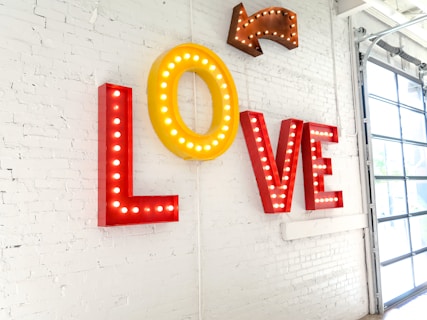 Large letters spelling out the word 'LOVE' are mounted on a white brick wall, with each letter brightly outlined by lightbulbs. An arrow with similar lighting points towards the word from above. The wall is part of a modern space with a large window partially visible on the right side.