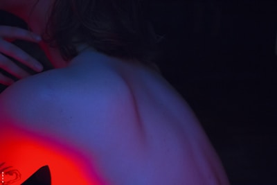 A close-up of a person's back with exposed skin, illuminated under a soft blue light. The lighting casts a red glow on the lower portion, adding an artistic contrast. A hand rests softly on the upper left, and part of a tattoo is visible in the red-tinted area.