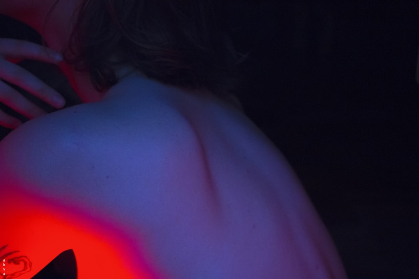 A close-up of a person's back with exposed skin, illuminated under a soft blue light. The lighting casts a red glow on the lower portion, adding an artistic contrast. A hand rests softly on the upper left, and part of a tattoo is visible in the red-tinted area.