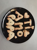 A black plate holds cookies shaped into letters and symbols, forming a message and including heart shapes. The cookies have a golden-brown color, typical of baked goods.