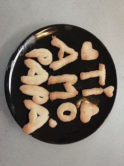 A black plate holds cookies shaped into letters and symbols, forming a message and including heart shapes. The cookies have a golden-brown color, typical of baked goods.