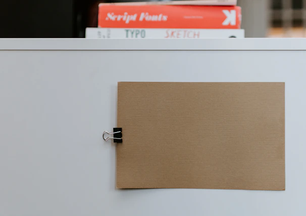 A beige piece of cardboard is clipped to a white board using a black binder clip. In the background, a stack of books features prominently, with visible titles including 'Script Fonts', 'TYPO', and 'SKETCH'. The arrangement appears organized and minimalist.