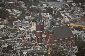 A densely packed neighborhood with rows of houses surrounding a large, prominent church. The church features tall spires, a large rose window, and intricate architectural details, standing out against the uniformity of nearby buildings. The area is characterized by a mix of trees and urban structures, creating a blend of natural and man-made elements.