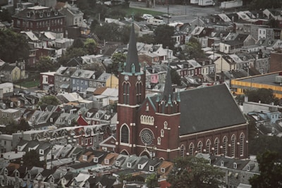 A densely packed neighborhood with rows of houses surrounding a large, prominent church. The church features tall spires, a large rose window, and intricate architectural details, standing out against the uniformity of nearby buildings. The area is characterized by a mix of trees and urban structures, creating a blend of natural and man-made elements.