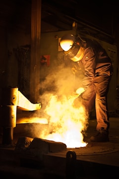A person wearing protective gear is working in an industrial setting with molten metal creating a bright glow. The surroundings are dimly lit, emphasizing the intense light and heat coming from the metal.