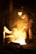 A person wearing protective gear is working in an industrial setting with molten metal creating a bright glow. The surroundings are dimly lit, emphasizing the intense light and heat coming from the metal.