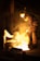 A person wearing protective gear is working in an industrial setting with molten metal creating a bright glow. The surroundings are dimly lit, emphasizing the intense light and heat coming from the metal.