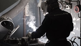 A person wearing protective gear, including a welding helmet and gloves, is engaged in welding in a workshop. Sparks and smoke are visible as bright white light emanates from the welding activity. The surroundings include various tools and equipment, such as a vent pipe, grinding wheels, and cords.