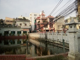 A small urban canal with a decorative stone bridge is bordered by residential and commercial buildings. The bridge features intricate stone carvings, and the canal is surrounded by greenery. Overhead, numerous black electrical wires create a complex, tangled pattern against the sky.