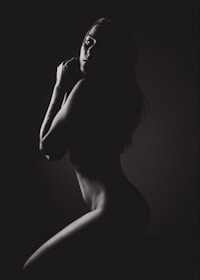 A monochrome image featuring the silhouette of a person in a side pose, with gentle light highlighting the contours of the face and body against a dark background.