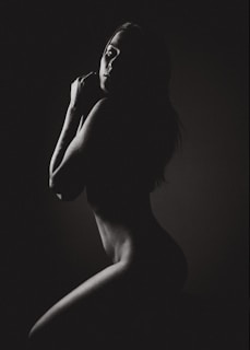 A monochrome image featuring the silhouette of a person in a side pose, with gentle light highlighting the contours of the face and body against a dark background.