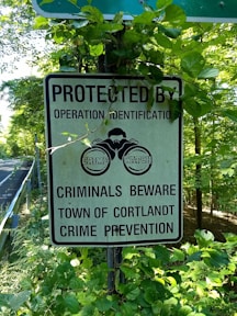 A sign is surrounded by green foliage on the side of a road. The sign reads 'Protected by Operation Identification. Criminals Beware. Town of Cortlandt Crime Prevention' and has an image of a pair of watchful eyes labeled 'Crime Watch'.