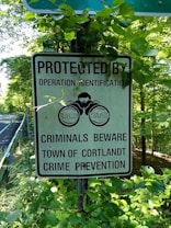 A sign is surrounded by green foliage on the side of a road. The sign reads 'Protected by Operation Identification. Criminals Beware. Town of Cortlandt Crime Prevention' and has an image of a pair of watchful eyes labeled 'Crime Watch'.