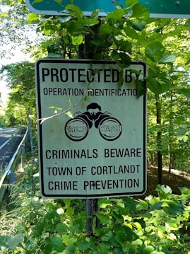 A sign is surrounded by green foliage on the side of a road. The sign reads 'Protected by Operation Identification. Criminals Beware. Town of Cortlandt Crime Prevention' and has an image of a pair of watchful eyes labeled 'Crime Watch'.