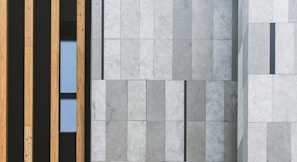A modern architectural facade featuring vertical wooden panels on the left side and large stone tiles with a subtle gradient on the right. A narrow window is placed between the wooden panels, adding a touch of contrast to the structured design.
