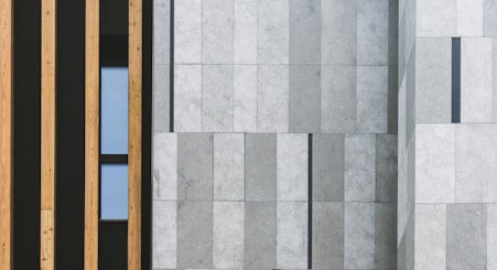 A modern architectural facade featuring vertical wooden panels on the left side and large stone tiles with a subtle gradient on the right. A narrow window is placed between the wooden panels, adding a touch of contrast to the structured design.