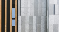 A modern architectural facade featuring vertical wooden panels on the left side and large stone tiles with a subtle gradient on the right. A narrow window is placed between the wooden panels, adding a touch of contrast to the structured design.