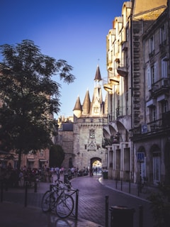 A cobblestone street lined with old architecture under a clear blue sky. The focal point is a historic stone gateway with conical turrets. People stroll down the street, adding to the lively atmosphere. Bicycles are parked in the foreground, and the buildings cast long shadows.
