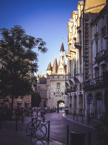 A cobblestone street lined with old architecture under a clear blue sky. The focal point is a historic stone gateway with conical turrets. People stroll down the street, adding to the lively atmosphere. Bicycles are parked in the foreground, and the buildings cast long shadows.