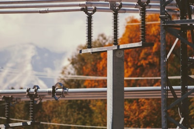 A close-up view of electrical infrastructure featuring metal poles and insulating coils. The background includes vibrant autumn foliage with shades of orange and red, and a faint outline of snow-capped mountains under a cloudy sky.