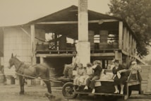 A rustic scene featuring a horse-drawn cart with people sitting on it. The cart is parked in front of a wooden building, likely used for industrial or agricultural purposes, with cylindrical structures and barrels visible. The setting appears rural and historical, with a group including children and adults casually sitting and chatting.