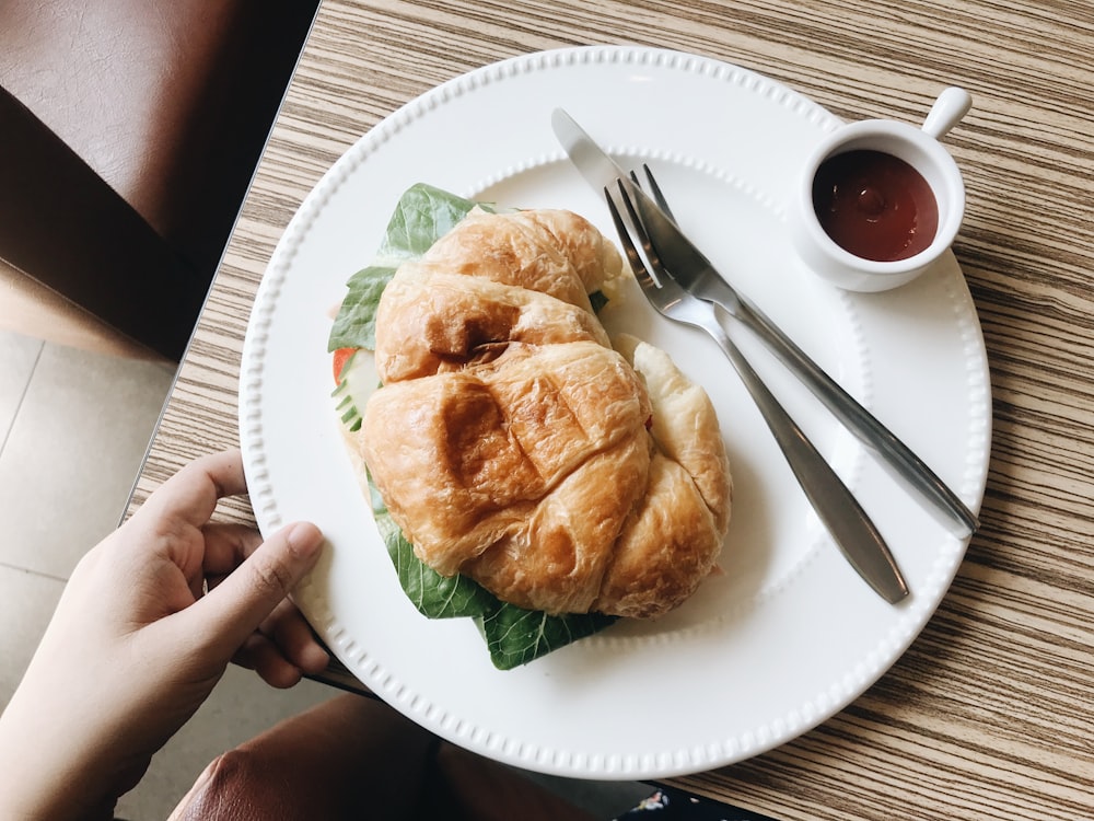 croissant on plate with spoon and fork and small white ceramic cup with sauce on table