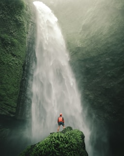 A person dressed in outdoor gear stands on a lush, green outcrop, gazing up at a massive waterfall cascading down a rugged cliff. Mist from the waterfall creates an ethereal atmosphere, surrounding the scene with a sense of grandeur and adventure.