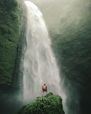 A person dressed in outdoor gear stands on a lush, green outcrop, gazing up at a massive waterfall cascading down a rugged cliff. Mist from the waterfall creates an ethereal atmosphere, surrounding the scene with a sense of grandeur and adventure.