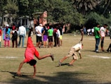 Two children are performing cartwheels on a grassy field, while a group of people stand in the background under the shade of trees. The scene is busy, suggesting a community event or gathering.