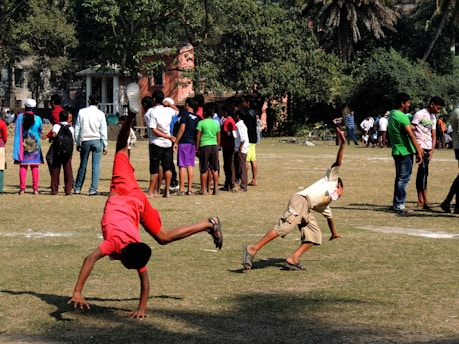 Two children are performing cartwheels on a grassy field, while a group of people stand in the background under the shade of trees. The scene is busy, suggesting a community event or gathering.