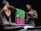 Two men are engaged in the process of dyeing fabric, holding a rod with vibrant pink and green textiles. The scene is dimly lit, highlighting their focused expressions and muscular builds. The fabric appears to be carefully draped over a rod, with one man on each side of the setup.