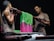 Two men are engaged in the process of dyeing fabric, holding a rod with vibrant pink and green textiles. The scene is dimly lit, highlighting their focused expressions and muscular builds. The fabric appears to be carefully draped over a rod, with one man on each side of the setup.