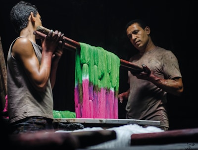 Two men are engaged in the process of dyeing fabric, holding a rod with vibrant pink and green textiles. The scene is dimly lit, highlighting their focused expressions and muscular builds. The fabric appears to be carefully draped over a rod, with one man on each side of the setup.