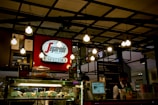 A coffee shop counter featuring a Segafredo Zanetti sign prominently displayed. The ceiling is adorned with multiple hanging light bulbs, creating a warm atmosphere. In the foreground, there is a glass display case filled with various pastries, and a person behind the counter appears to be serving customers.