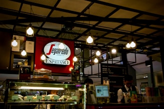 A coffee shop counter featuring a Segafredo Zanetti sign prominently displayed. The ceiling is adorned with multiple hanging light bulbs, creating a warm atmosphere. In the foreground, there is a glass display case filled with various pastries, and a person behind the counter appears to be serving customers.