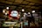 A coffee shop counter featuring a Segafredo Zanetti sign prominently displayed. The ceiling is adorned with multiple hanging light bulbs, creating a warm atmosphere. In the foreground, there is a glass display case filled with various pastries, and a person behind the counter appears to be serving customers.