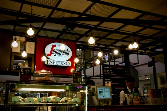 A coffee shop counter featuring a Segafredo Zanetti sign prominently displayed. The ceiling is adorned with multiple hanging light bulbs, creating a warm atmosphere. In the foreground, there is a glass display case filled with various pastries, and a person behind the counter appears to be serving customers.