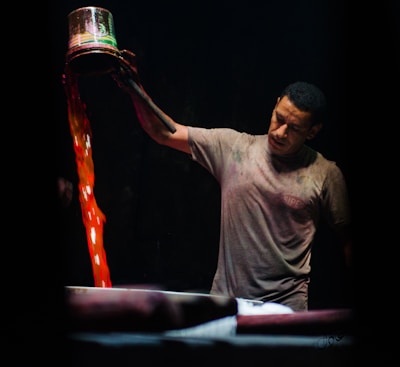 A man is pouring liquid from a metal container with vibrant red and orange hues into a large vat. He appears to be focused and intent on his task. The background is dark, highlighting both the man and the vivid colors of the liquid.