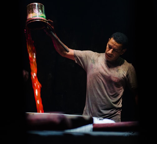 A man is pouring liquid from a metal container with vibrant red and orange hues into a large vat. He appears to be focused and intent on his task. The background is dark, highlighting both the man and the vivid colors of the liquid.