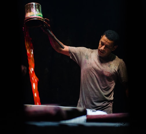 A man is pouring liquid from a metal container with vibrant red and orange hues into a large vat. He appears to be focused and intent on his task. The background is dark, highlighting both the man and the vivid colors of the liquid.